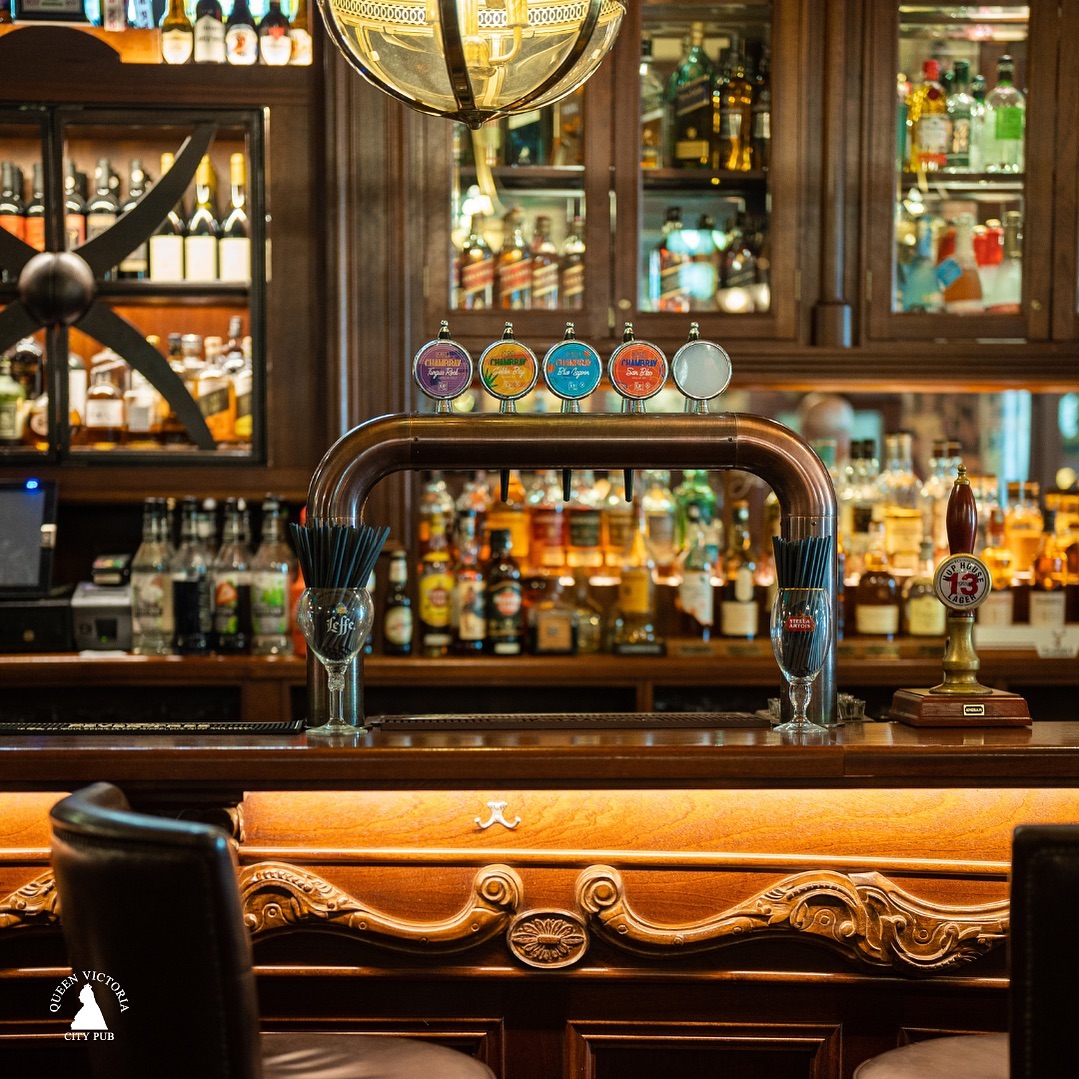 Draft beer taps and liquor bottles lined up at the wooden bar counter inside The Queen Victoria City Pub