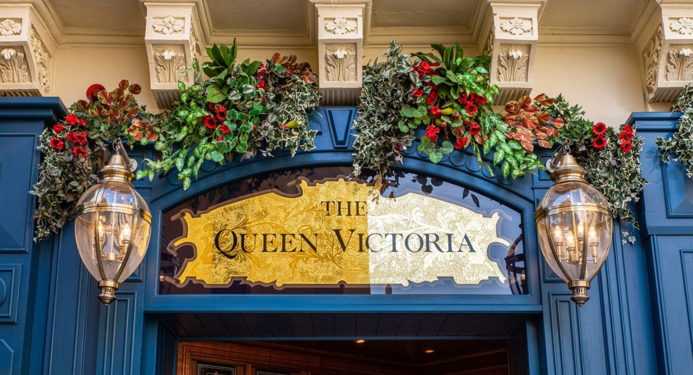 Gold vintage glass signboard with flower garlands and brass lamps above the entrance of The Queen Victoria City Pub